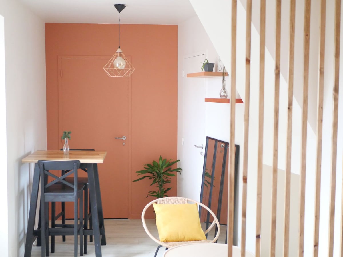 A corner of the studio features a wooden bar table paired with tall stools, complemented by a small table and chair. A potted plant adds a touch of greenery. An accent wall in orange provides a warm backdrop, while light fixtures and decorative shelves enhance the space.
