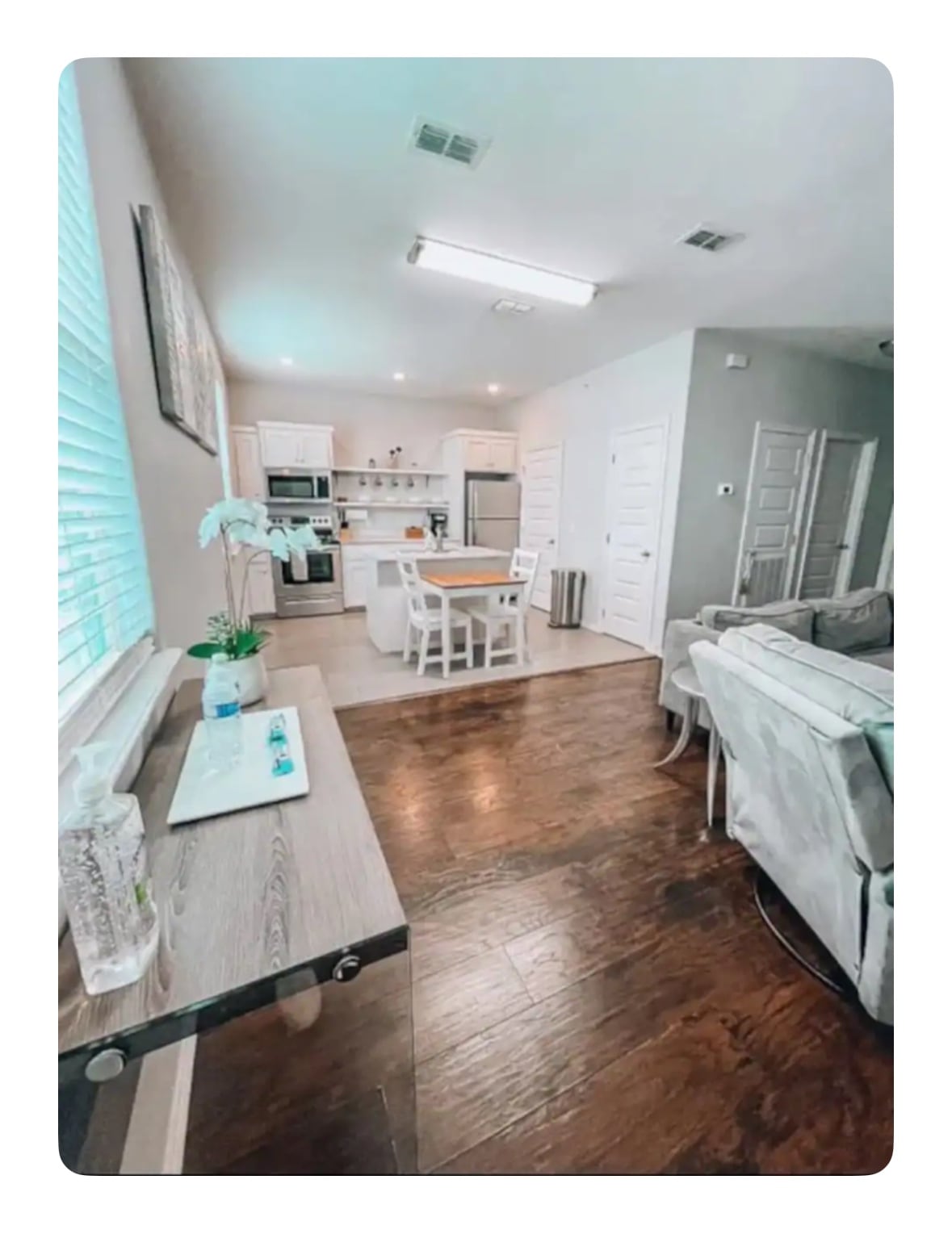 A spacious living area connects to a modern kitchen, provided with a wooden table surrounded by white chairs. Light-colored cabinets and appliances create a bright ambiance. A small side table with decorative items enhances the room's functionality. Large windows allow natural light to brighten the space.