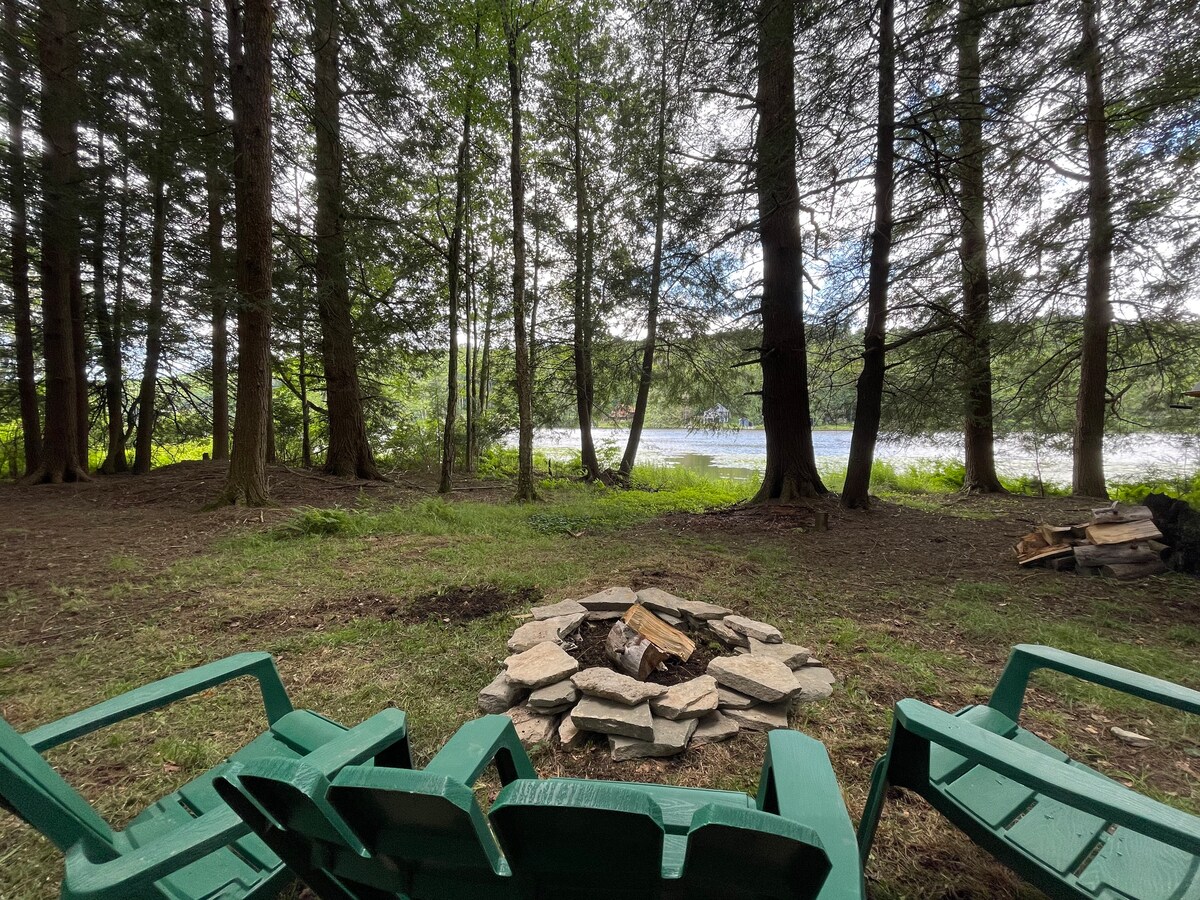 A circular stone fire pit is surrounded by green adirondack chairs, set in a clearing surrounded by tall trees. Beyond the tree line, a serene water view is visible, with sunlight filtering through the branches, creating a peaceful atmosphere.