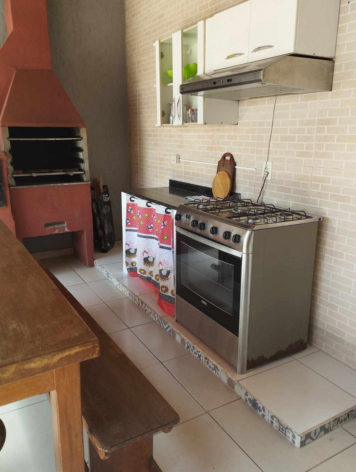 A well-equipped kitchen area features a gas stove, an oven, and a kitchen exhaust hood. A colorful dish towel adorns the stove. A rustic wooden table provides space for dining, and a brick barbecue grill is visible nearby, complementing the functional design.