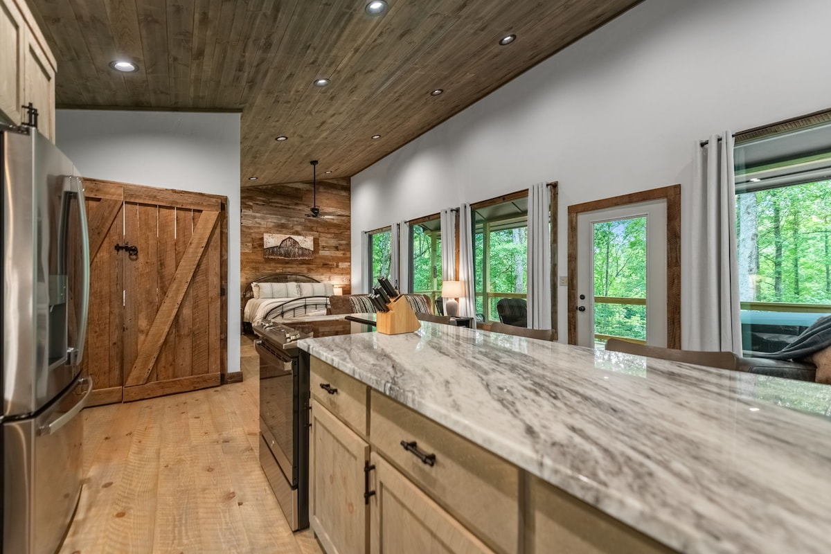 A spacious kitchen and living area is highlighted by natural wood accents and contemporary furnishings. The marble countertop contrasts with rustic cabinetry, and large windows allow natural light to fill the space, showcasing views of the lush greenery outside.