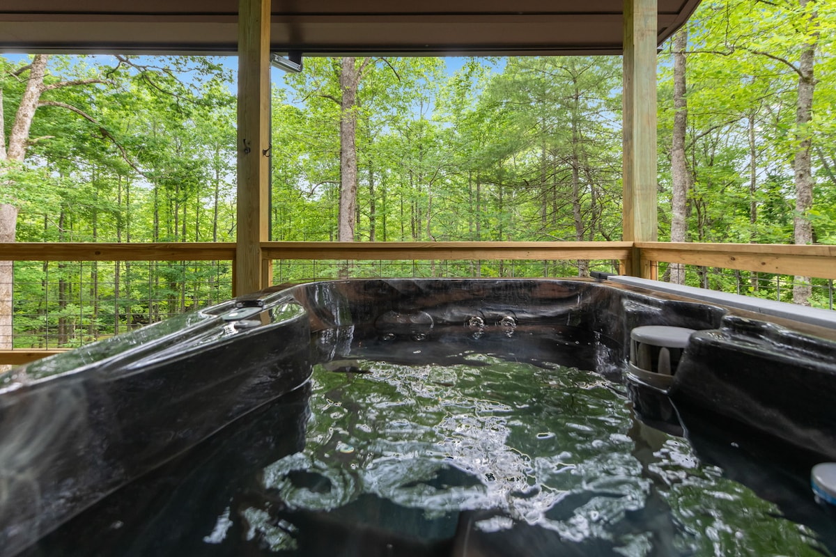 A hot tub filled with clear water is positioned on a deck, surrounded by lush green trees. The natural setting creates an atmosphere of privacy and tranquility, while the wooden railing offers a glimpse of the forested landscape beyond.