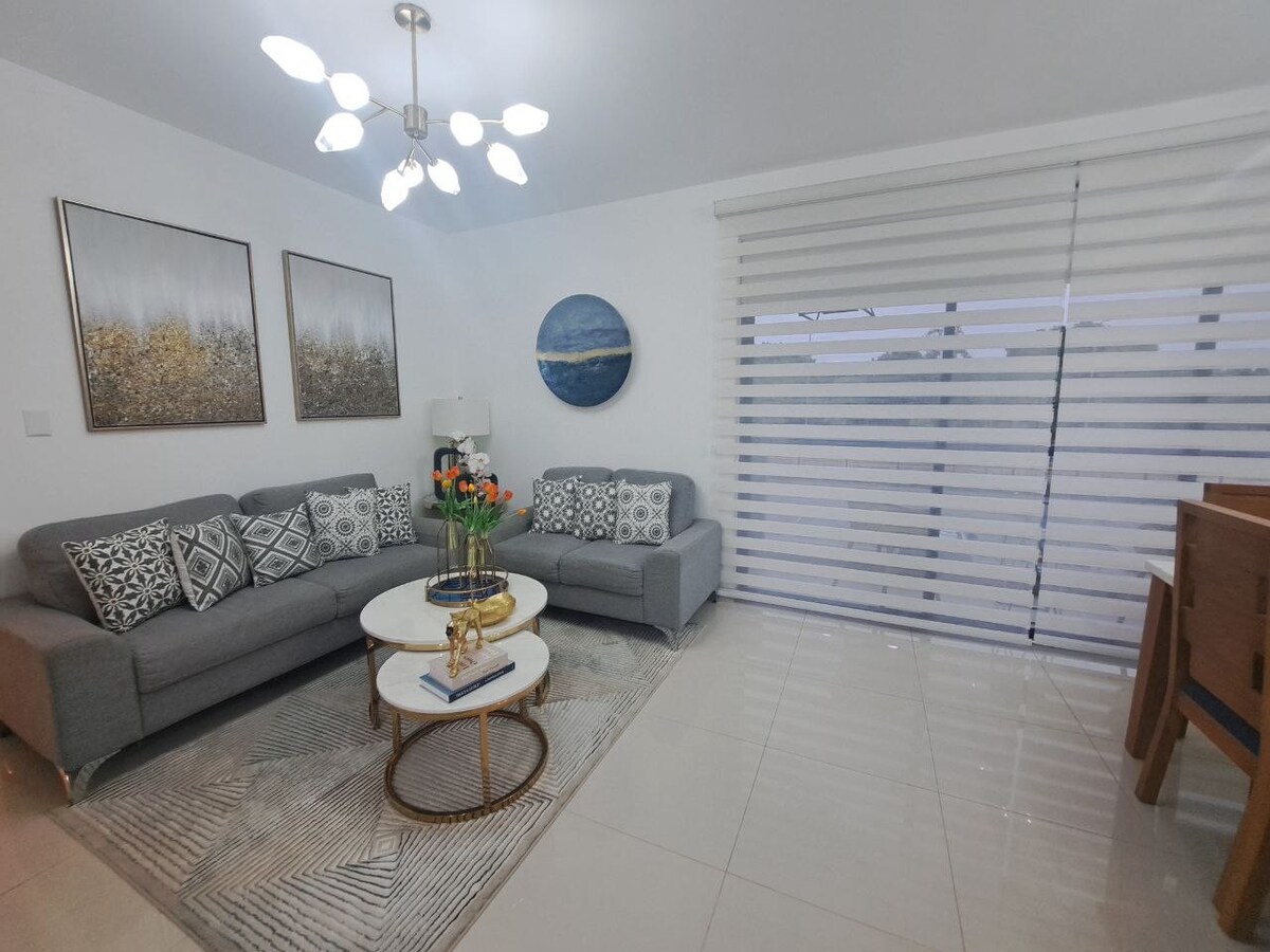 A modern living area is furnished with two gray sofas adorned with decorative pillows. An abstract artwork is displayed on the walls, complemented by a round coffee table. Natural light filters through elegantly striped window coverings, creating a bright and open ambiance.