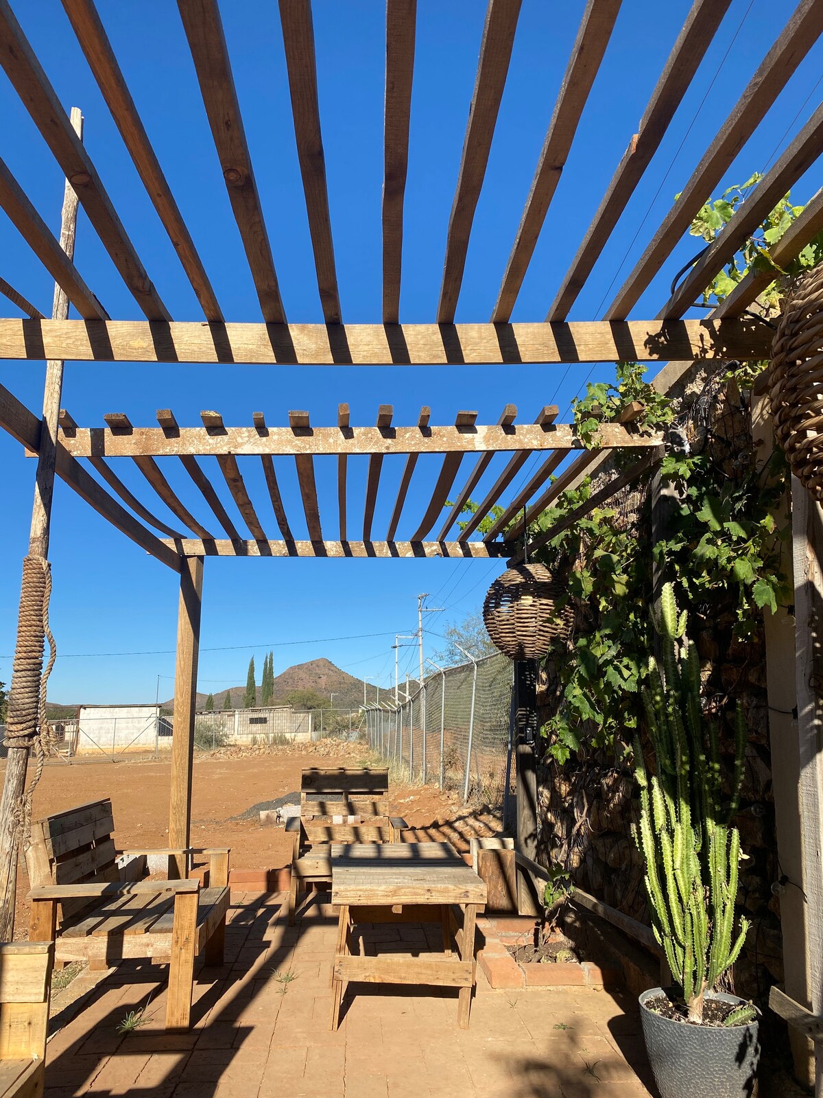 An outdoor seating area is framed by wooden beams, providing shade. Natural materials are used for the furniture, and potted cacti add greenery. The space is surrounded by the open landscape, with a clear blue sky visible above.