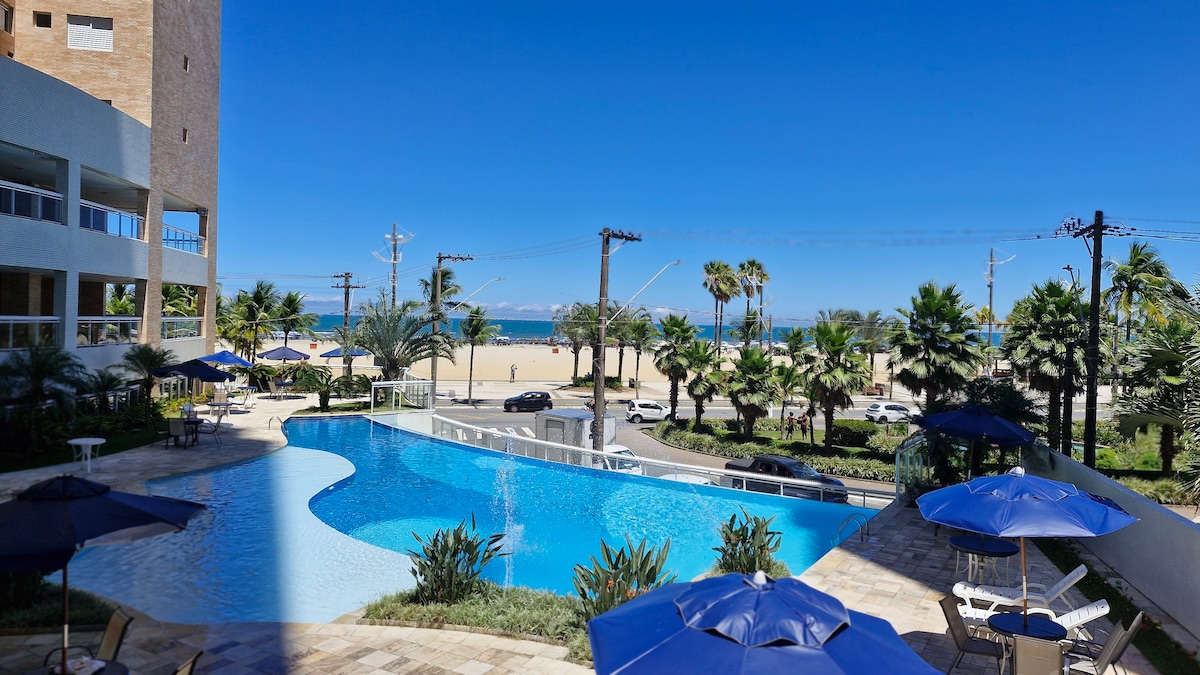 An expansive outdoor infinity pool is visible, surrounded by lush greenery and lounge chairs. Umbrellas provide shade for guests, while the beautiful ocean and sandy beach can be seen in the background under a clear blue sky.