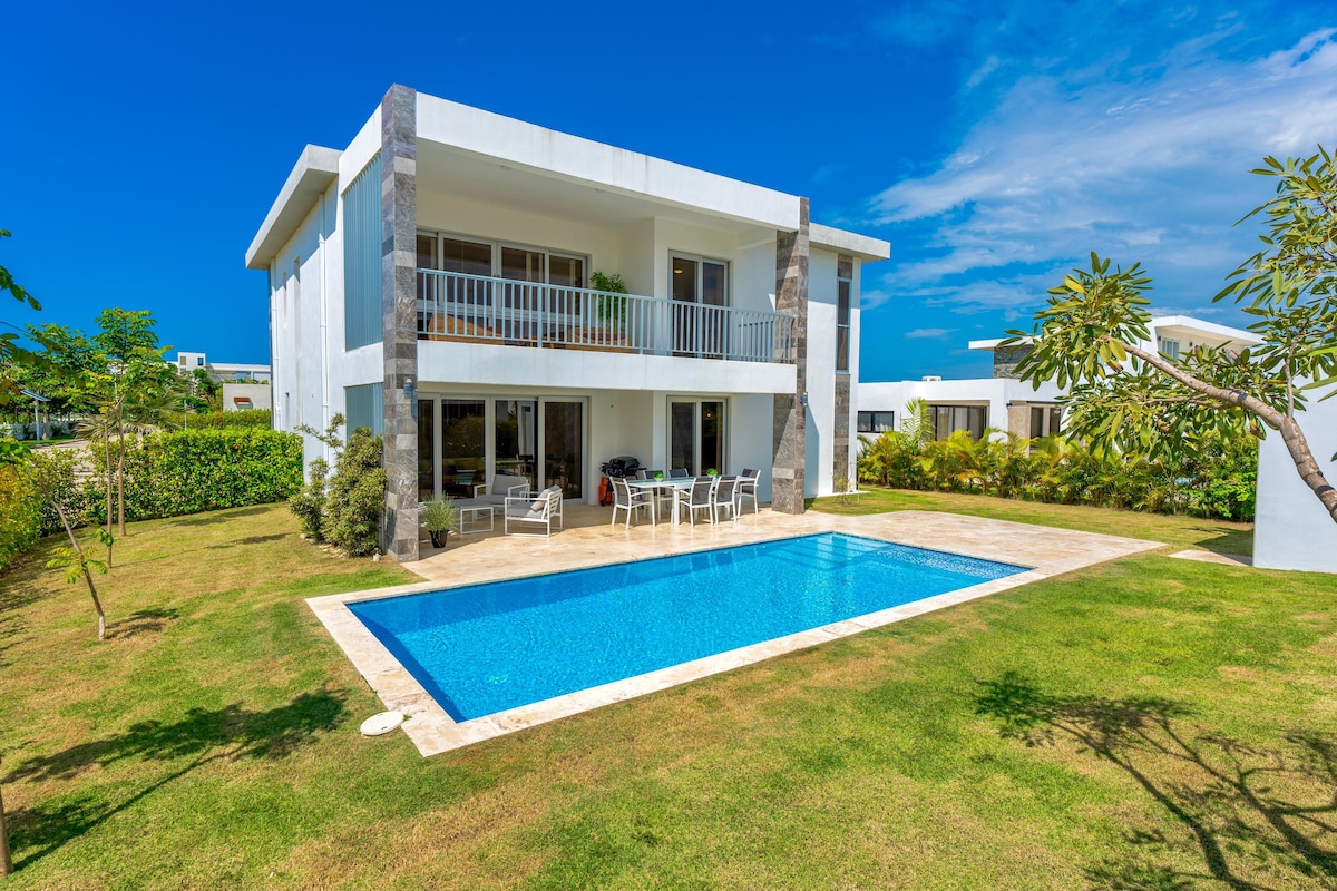 A modern two-story villa is surrounded by lush greenery, featuring a sparkling blue pool in the backyard. The spacious terrace includes several white chairs and a table, offering a space for relaxation and outdoor dining. Large glass doors provide views of the interior.
