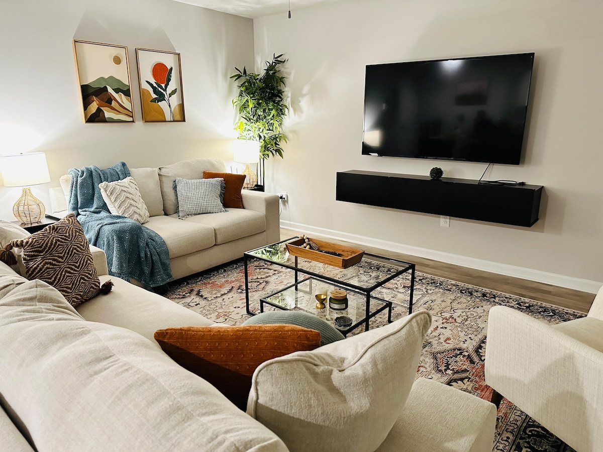 A comfortable living area features two soft sofas arranged around a glass coffee table. A large wall-mounted TV is prominently displayed, and a floor lamp provides additional lighting. Decorative artwork and a green plant add warmth to the neutral-toned space.