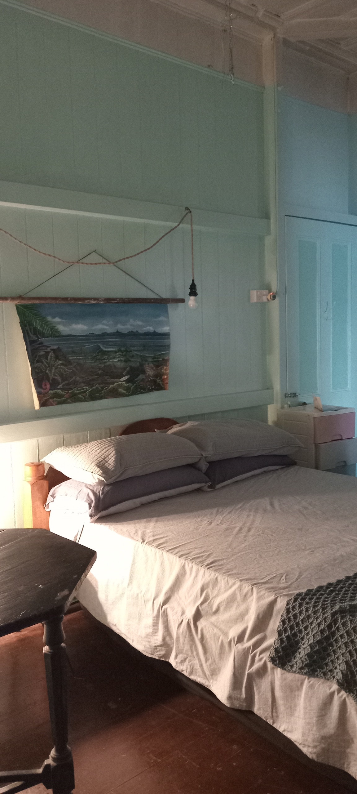 A cozy sleeping area features a spacious bed dressed in soft gray linens, complemented by plump pillows. A colorful landscape painting hangs on the light blue wall, while a small side table provides a surface for personal items. Natural light gently illuminates the room.