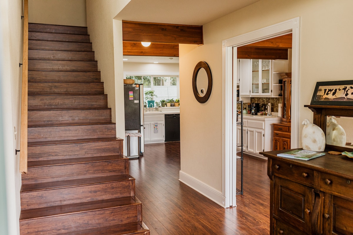 A spacious entryway is highlighted by a wooden staircase leading to the upper level. Natural light filters through large windows, illuminating the open-plan layout that connects the kitchen and living areas. A round mirror adorns the wall, reflecting the inviting ambiance.