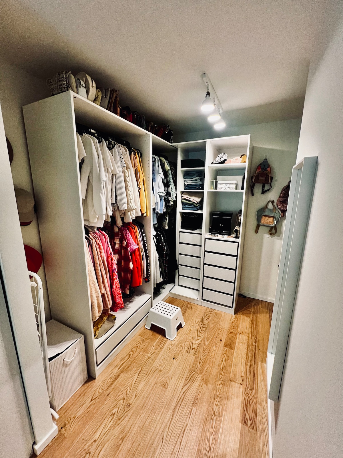 A well-organized closet space is displayed, featuring a combination of hanging clothing and neatly arranged shelves. A small step stool is positioned on the floor, and a full-length mirror is visible on the left side, reflecting the room's light wood flooring.