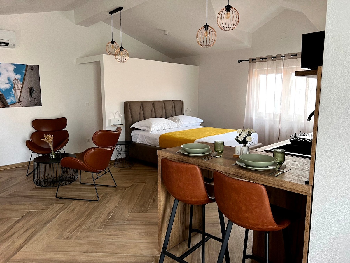 A spacious studio is shown, featuring a comfortable bed with crisp white linens and a warm yellow throw. Two modern armchairs sit across from a wooden dining table set for two, complemented by stylish lighting fixtures and large windows allowing natural light to fill the room.