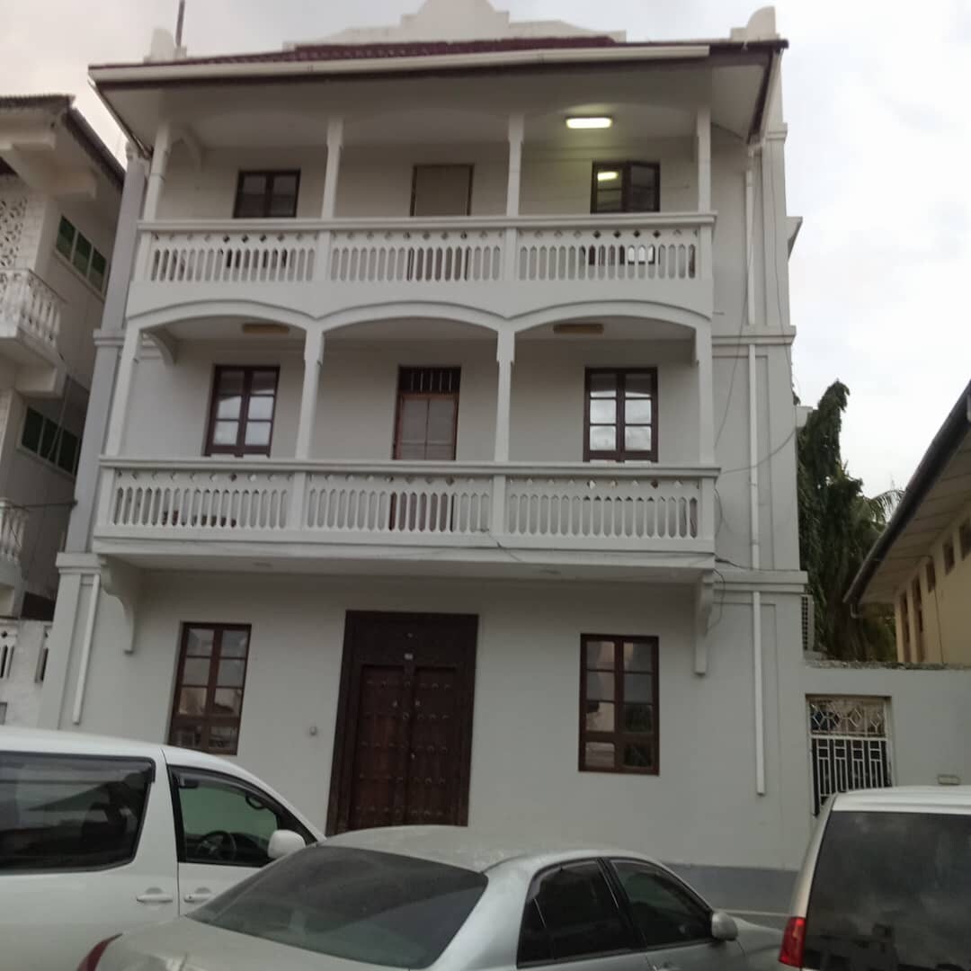 The exterior of a three-story building is displayed, featuring a light-colored facade with white balconies. Multiple windows are visible, framed in brown. A wooden door is centered on the ground level, with vehicles parked in front and surrounding the property.