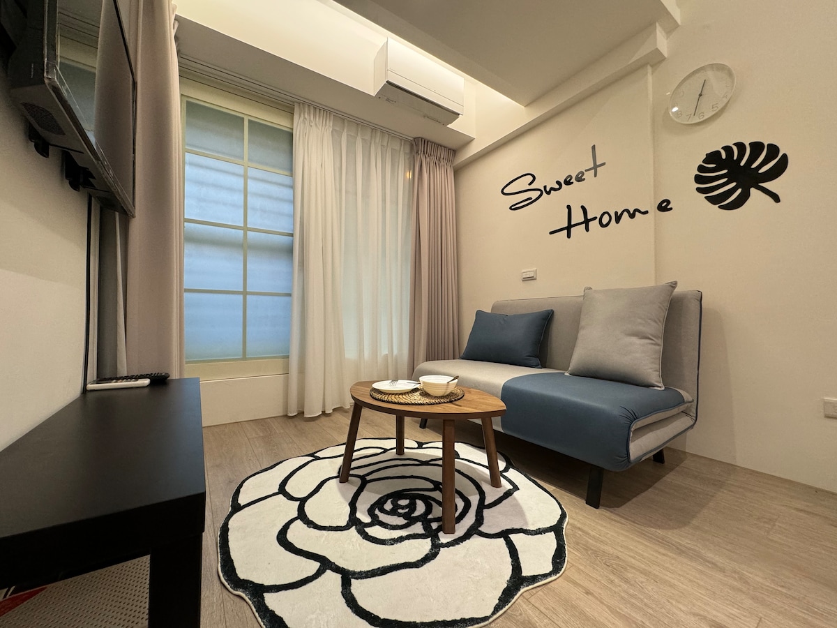 A cozy corner of the room features a light blue sofa against a wall with a decorative mural reading 'Sweet Home.' A round table with plates sits on a floral rug, while soft curtains allow natural light to filter in through the window.