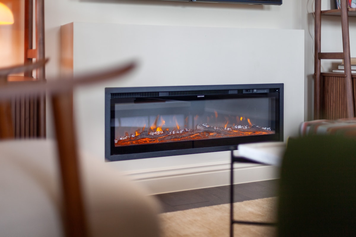 A modern linear fireplace is centrally positioned along a smooth wall, with glowing embers visible beneath a glass front. The surrounding area features neutral tones and complementary furnishings, enhancing the cozy ambiance of the living space.