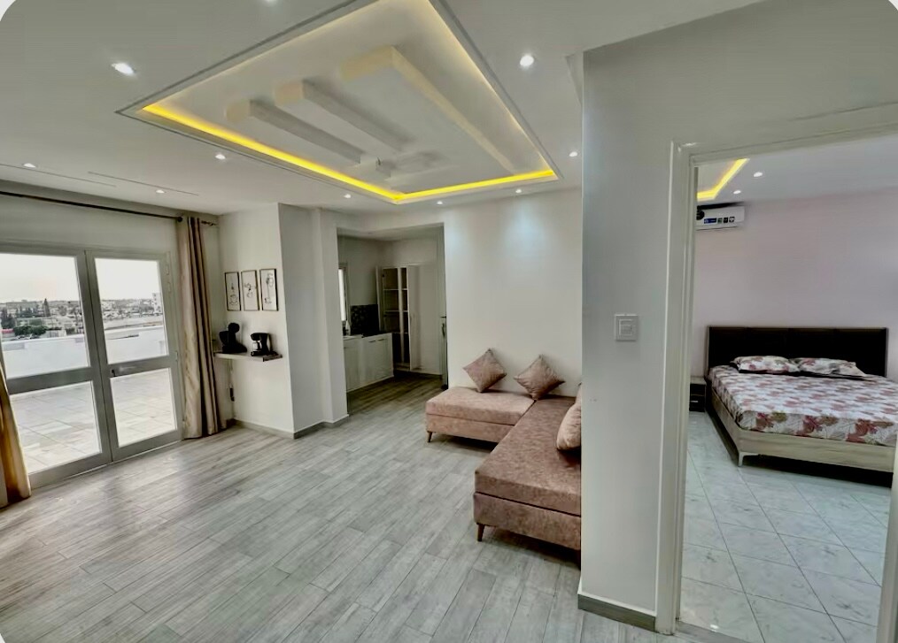 An open living area features a comfortable seating arrangement alongside large windows allowing natural light. A modern kitchenette can be seen in the background. A separate sleeping area is accessible, showcasing a spacious bed adorned with floral bedding.