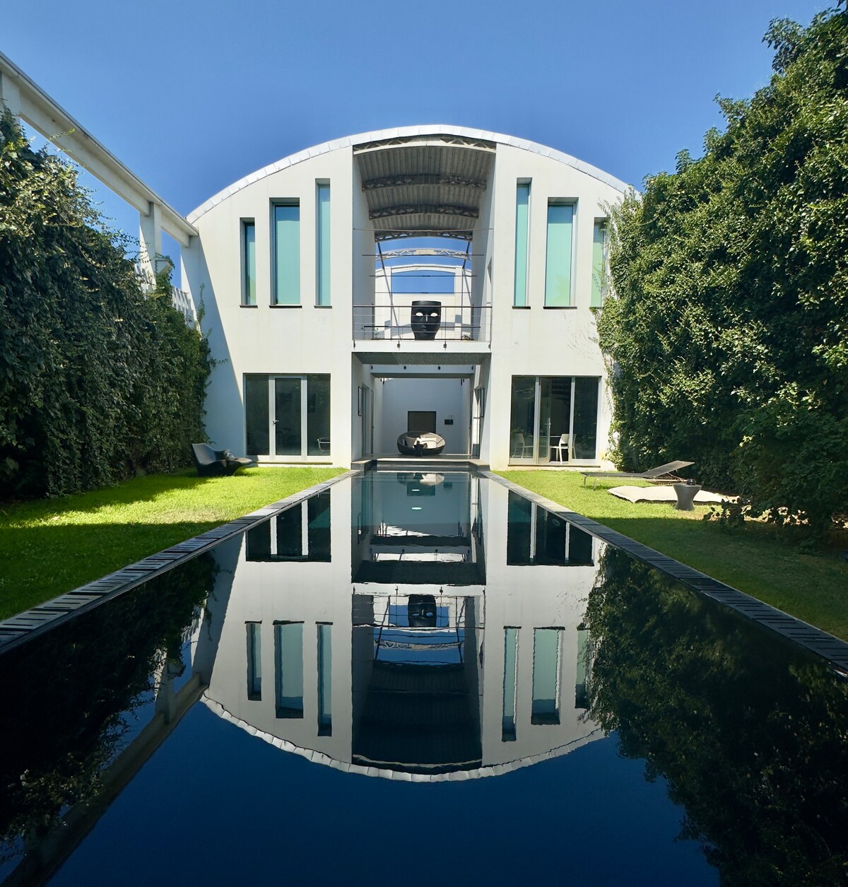 A modern building is mirrored in a calm reflecting pool, surrounded by well-maintained greenery. Large vertical windows allow natural light to enter, while a minimalistic facade features a distinct architectural design that draws attention.