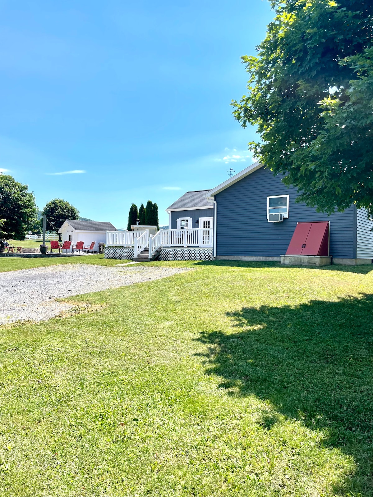 The exterior of the guest house is shown, featuring a grey facade complemented by white trim. A covered porch is visible, and a red door adds a pop of color. The surrounding lush green lawn and clear blue sky create a serene outdoor setting.