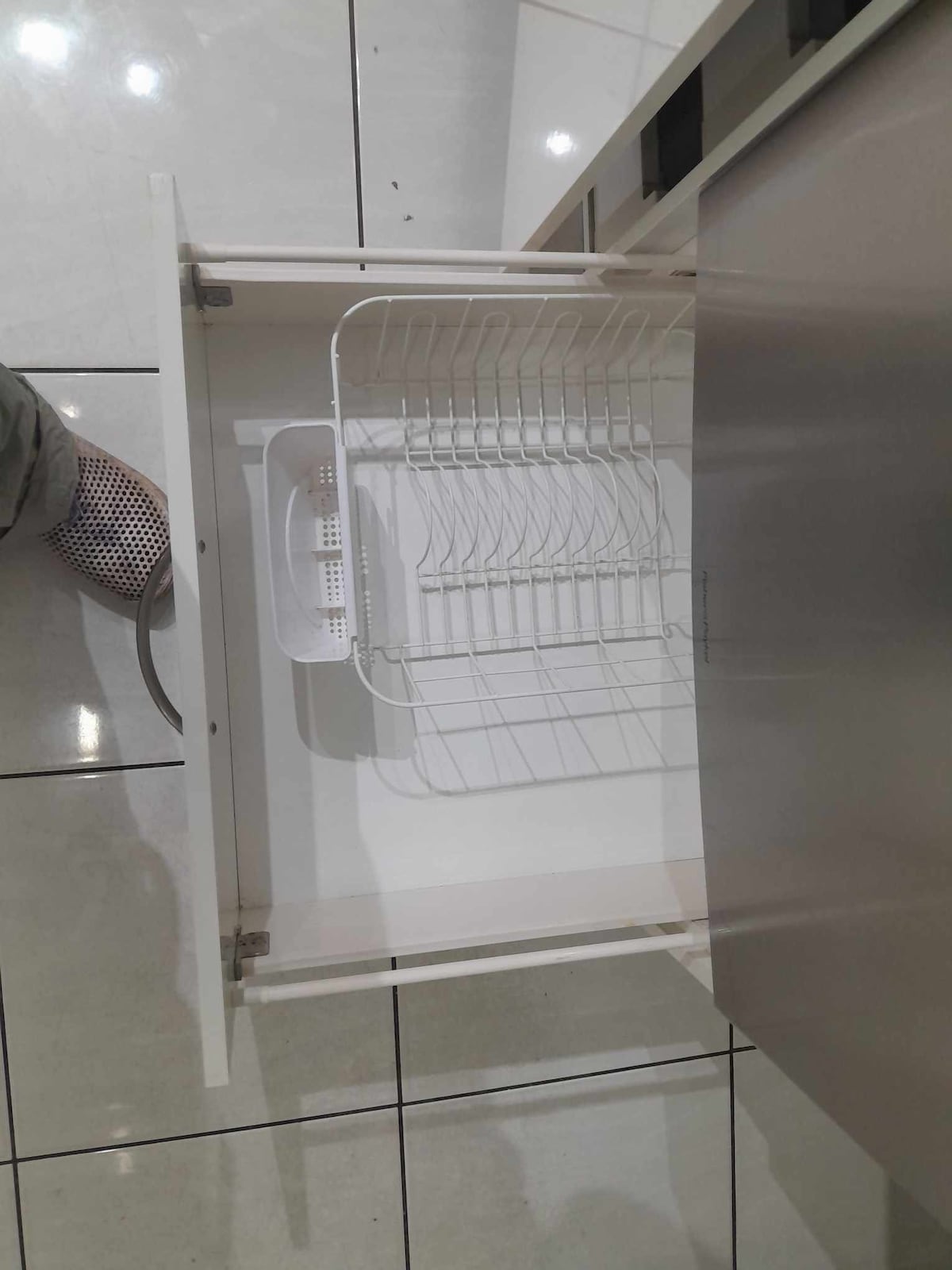 An open kitchen drawer is presented, featuring a removable wire rack and a small plastic container. The drawer’s interior is clean and spacious, with a smooth white finish and reflective tiled flooring visible in the image.
