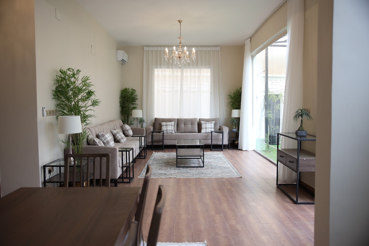 A spacious living area features comfortable seating arrangements with decorative cushions. Large windows allow natural light to fill the room, while a modern chandelier hangs above. Potted plants add a touch of greenery, and a rug complements the wooden flooring.