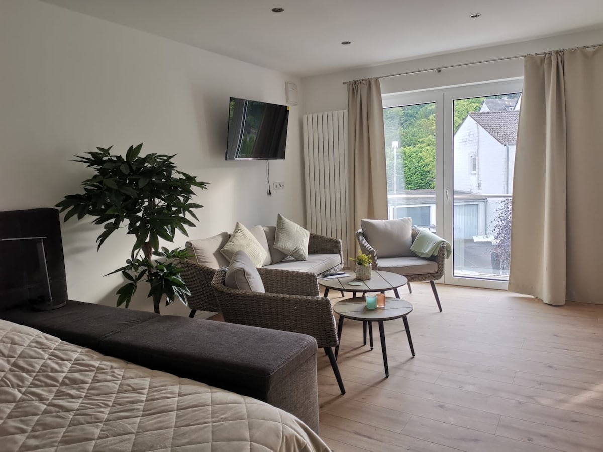 A cozy living area showcases a comfortable seating arrangement with two chairs and a side table. Natural light enters through large glass doors, enhancing the neutral tones of the decor. A plant adds a touch of greenery, contributing to a bright and inviting atmosphere.