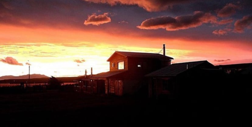 A serene outdoor scene captures a silhouette of a two-story cabin against a vibrant sunset. The sky displays a gradient of oranges, pinks, and purples, providing a natural backdrop. The overall setting suggests a peaceful, rural environment, ideal for relaxation and connection with nature.