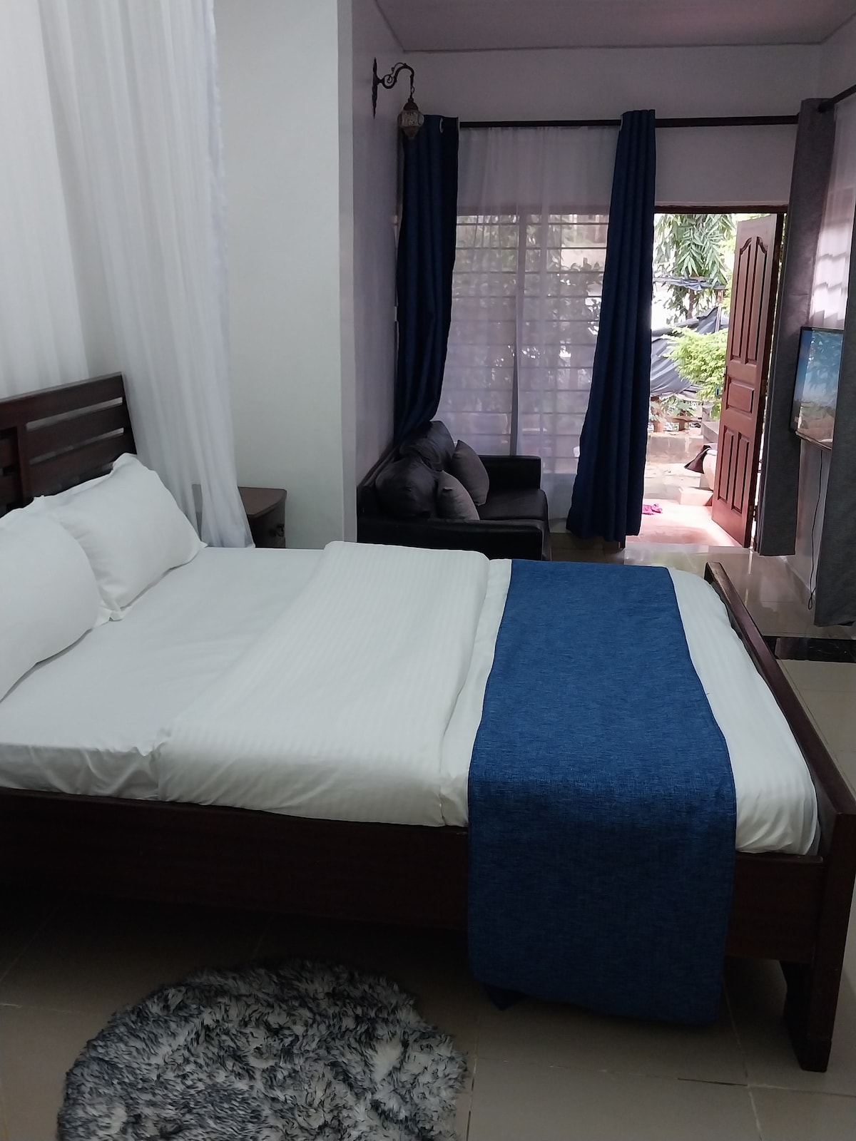 A cozy bedroom features a spacious bed dressed in crisp white linens, topped with a blue throw blanket. Natural light filters through sheer curtains, illuminating the room. A small seating area is visible near the entrance, enhancing the inviting atmosphere.