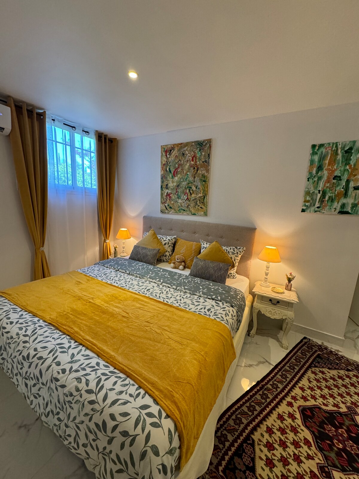 A cozy bedroom is featured with a large bed layered with an orange throw and patterned bedding. Two bedside lamps provide soft lighting, while artistic decorations adorn the walls. A window, framed by curtains, allows natural light to brighten the space.
