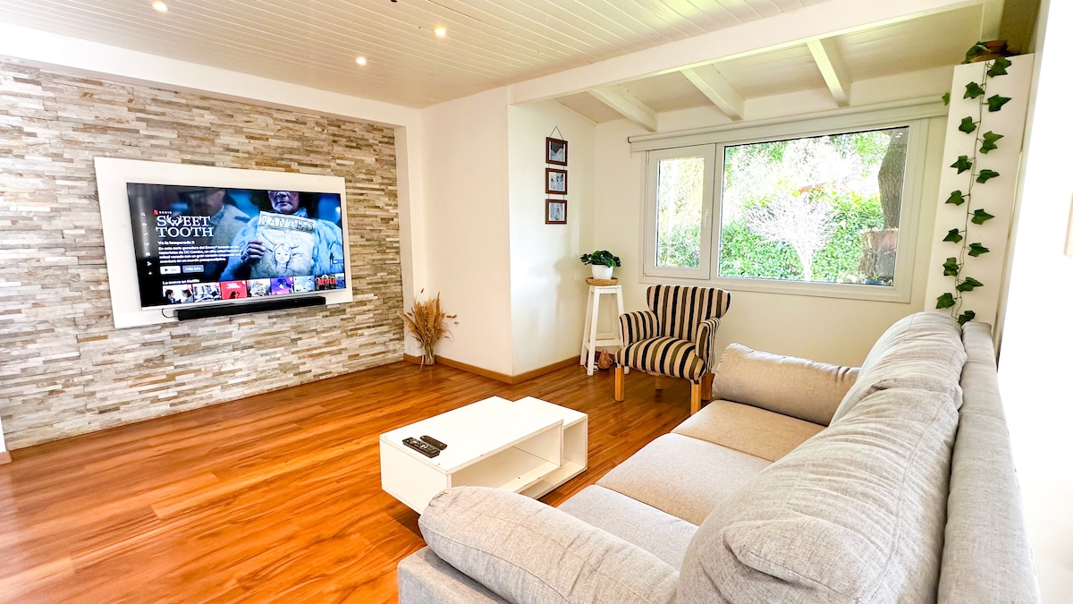 A bright living area is presented, featuring a large wall-mounted television and a cozy sectional sofa positioned for relaxation. Natural light enters through a sizable window, framed by greenery. A small white coffee table adds a functional touch, complemented by a striped armchair in the corner.