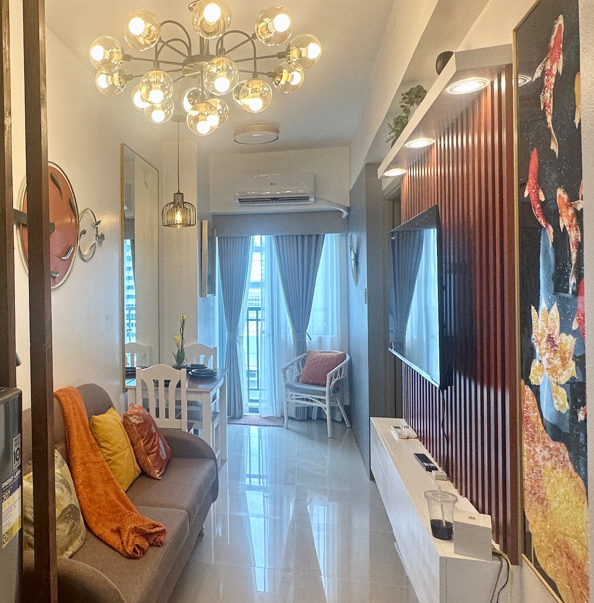 A well-lit living area features a three-seater sofa adorned with orange throws, complemented by a stylish chandelier overhead. A television is mounted on the wall, surrounded by decorative panels. Natural light flows through the sheer curtains over the balcony door, enhancing the cozy ambiance.