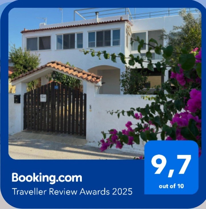 A charming villa exterior is framed by vibrant bougainvillea flowers. The whitewashed walls and tiled roof create an inviting entrance, with a wooden gate leading to the property. Natural light brightens the scene, highlighting the welcoming atmosphere of the home.