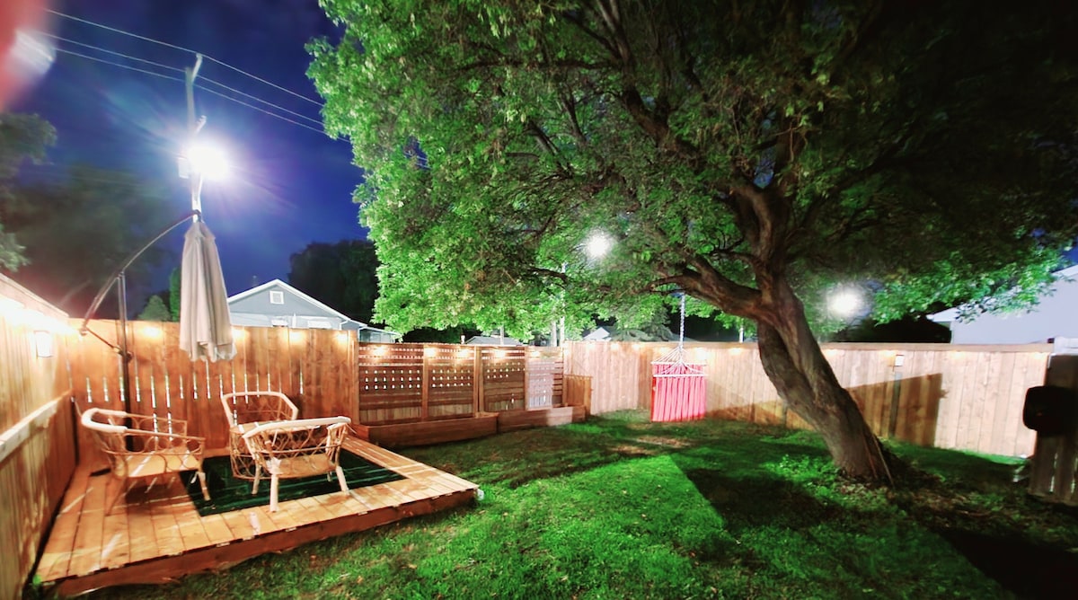 A spacious outdoor area features a grassy yard illuminated by soft lighting. A large tree provides shade over a wooden deck equipped with chairs. A privacy fence surrounds the space, ensuring a secluded atmosphere for relaxation.