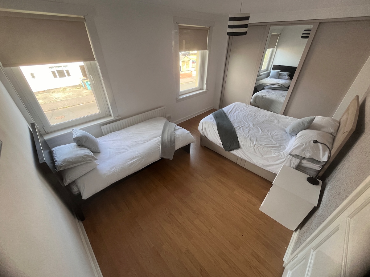Two bedrooms are visible, featuring a spacious bed and a single bed, both adorned with simple linens. A mirrored wardrobe reflects natural light from the windows, enhancing the room's brightness. The hardwood floor adds warmth, while minimalistic lamps and a small nightstand provide functionality.