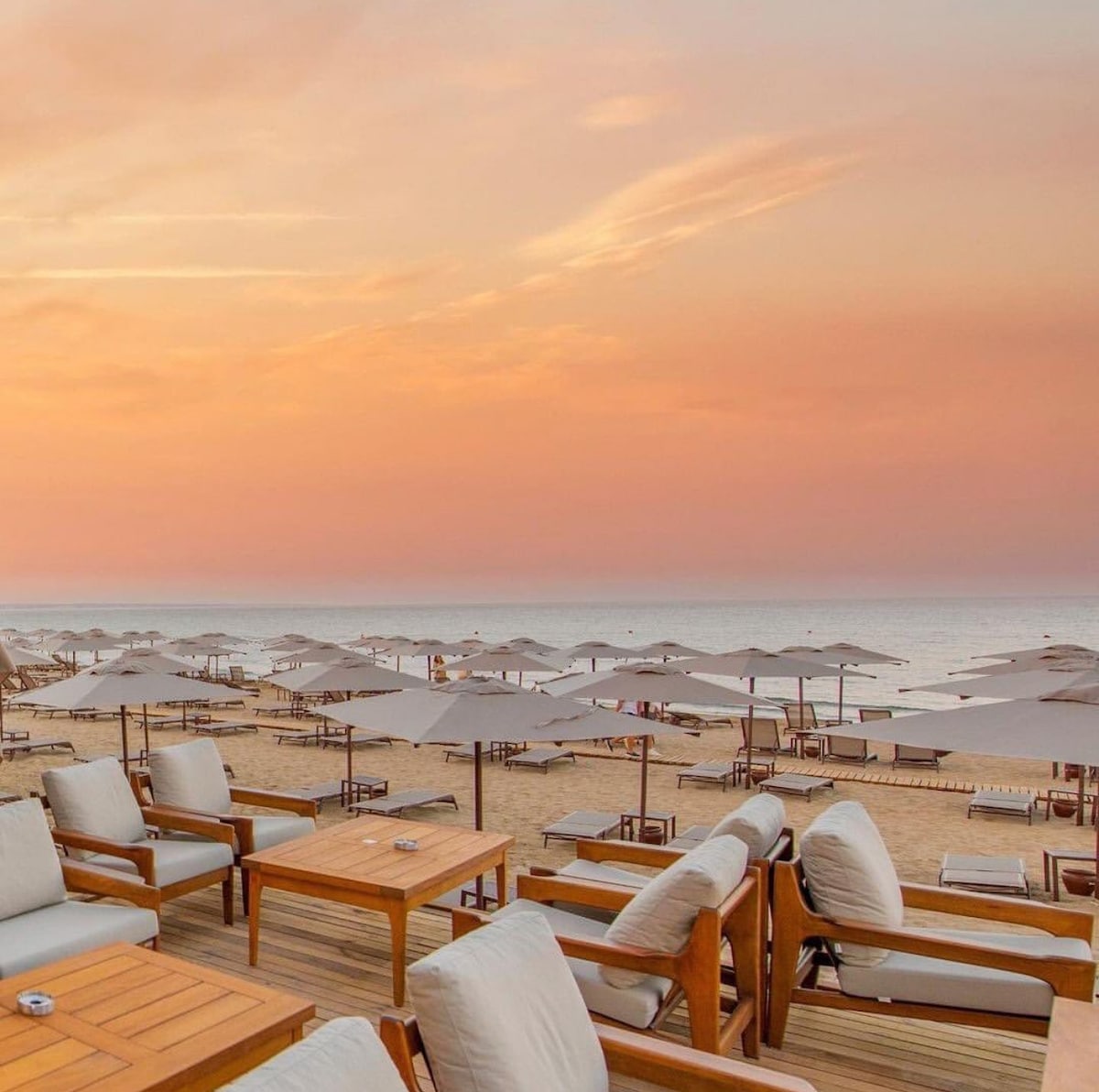 A serene beachfront view captures comfortable lounge chairs surrounding wooden tables on a sandy surface. Umbrellas are positioned to provide shade, with the ocean gently lapping at the shore. The sky is painted in soft hues of orange and pink, reflecting a tranquil atmosphere.