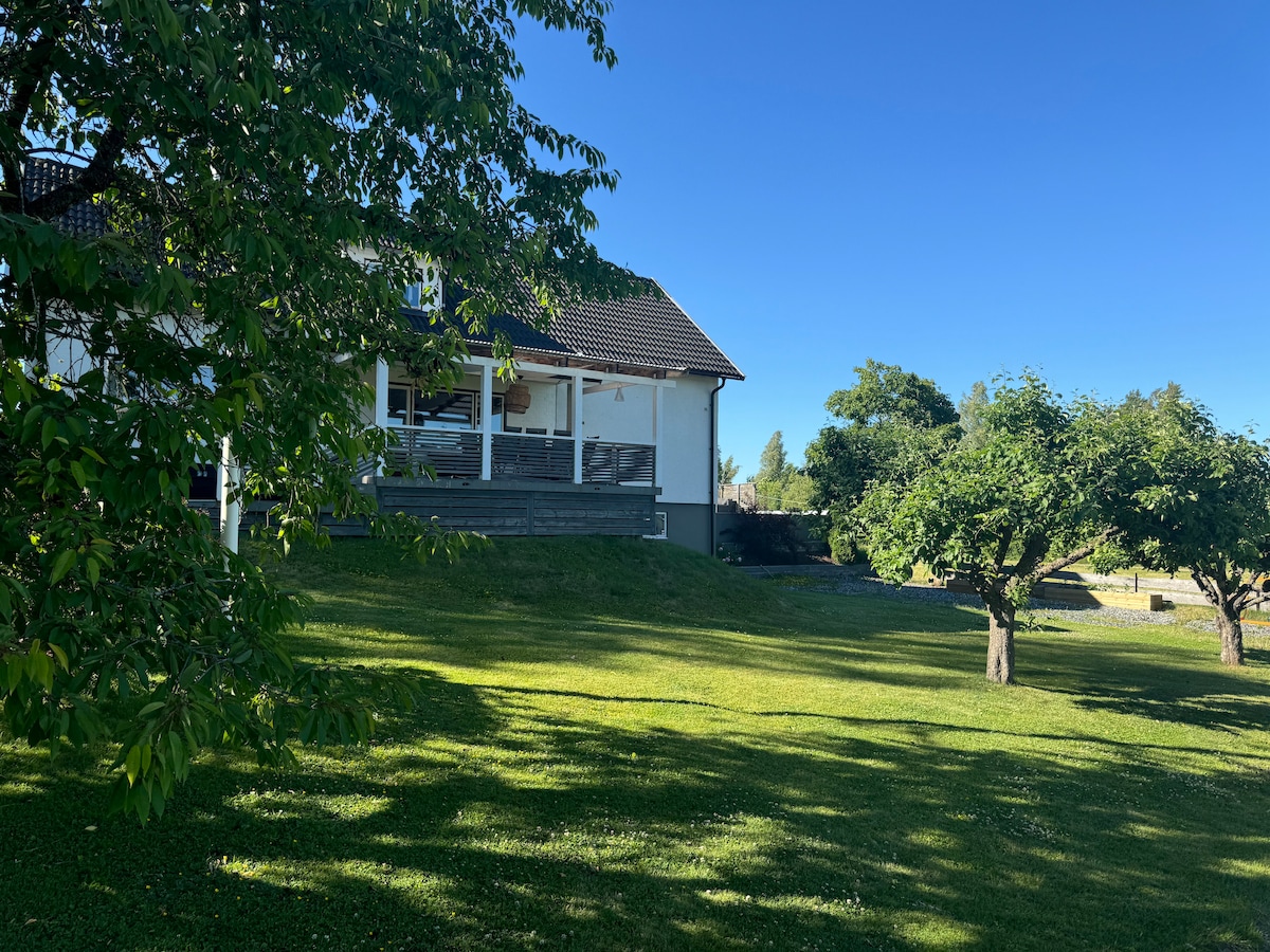 The exterior of the modern villa is visible, framed by lush green grass and surrounding trees. A large patio area is seen, providing space for outdoor relaxation. The clear blue sky creates a serene backdrop, emphasizing the peaceful setting.