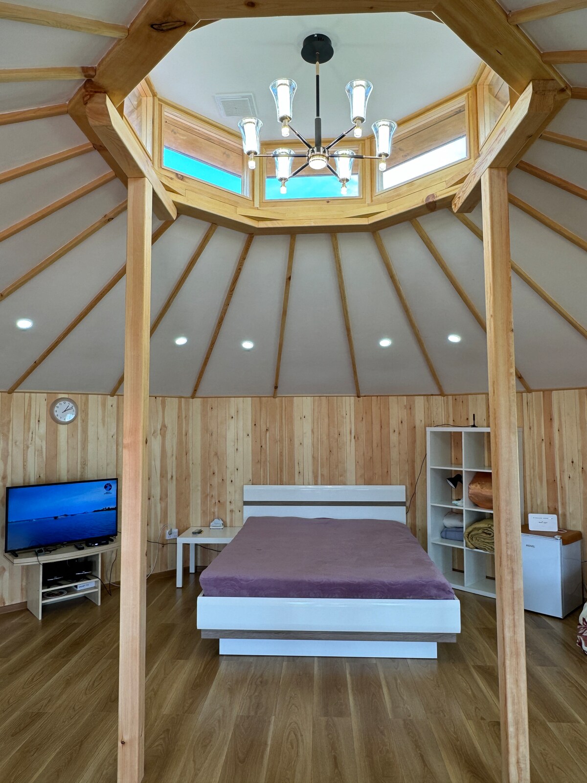 The interior of the Ger features wooden walls and a circular skylight, providing natural light. A spacious bed with a purple coverlet is centered in the room. A TV and a shelf with decorative items are positioned nearby, contributing to the cozy atmosphere.