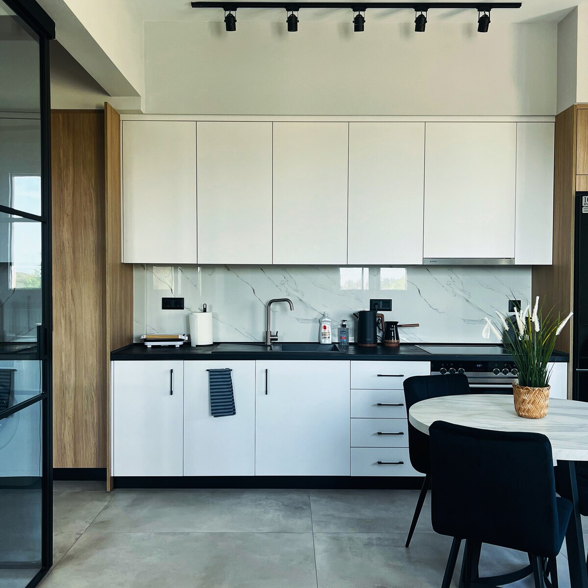 A modern kitchen area features sleek white cabinets and a dark countertop. A small dining table is set with black chairs, and a subtle vase holds fresh greenery. Natural light filters through a nearby window, enhancing the clean aesthetic of the space.