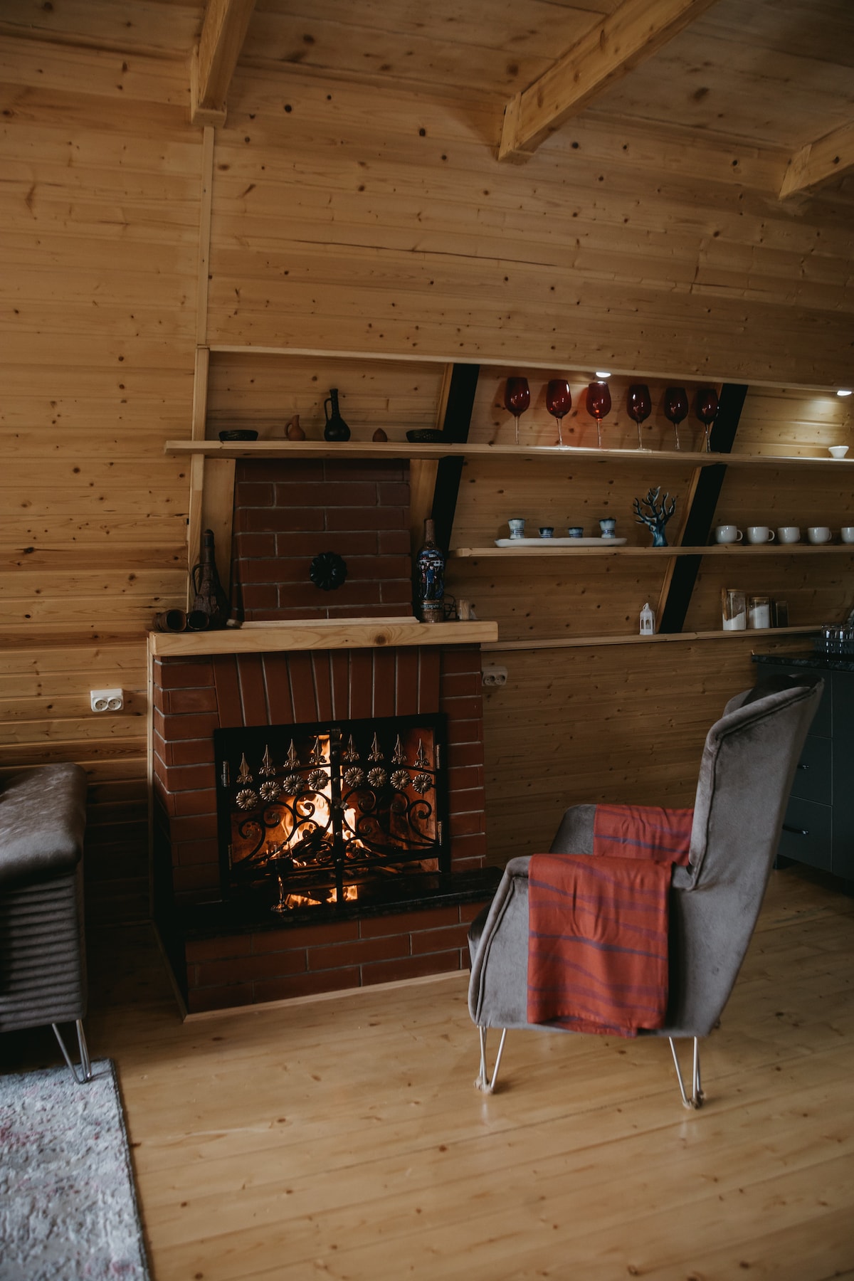 A cozy seating area features a brick fireplace with a decorative metal grate, flickering flames visible inside. Warm wooden walls and shelves display various items. A plush armchair, draped with a red throw, is positioned for relaxation in this inviting space.