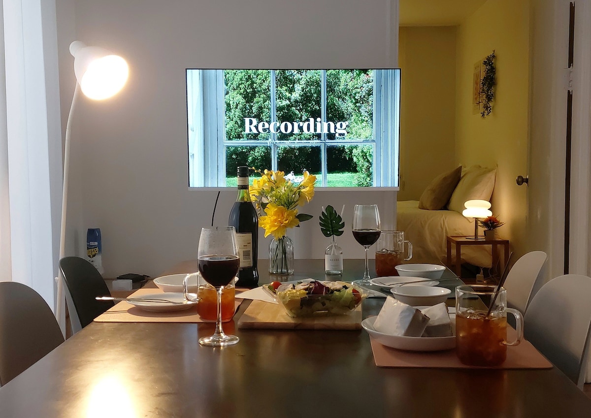 A dining table is set for a meal, featuring beautifully arranged plates and drinks. A modern flat-screen TV displays the word 'Recording' framed by a window view of greenery. A warm lamp and decorative vase with yellow flowers complement the inviting space.