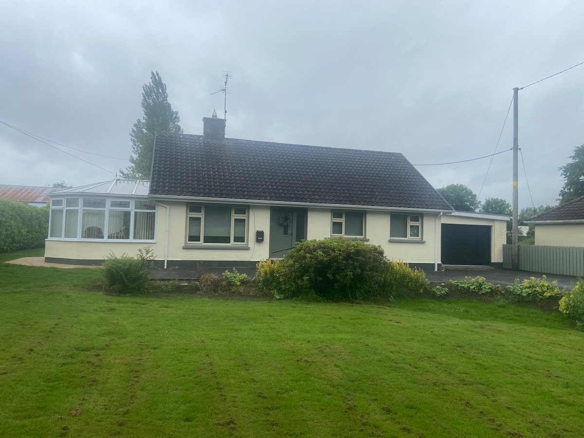 A single-story cottage is set against a cloudy sky, featuring a landscaped front yard with an expanse of grass and surrounding greenery. A conservatory extends from the front of the cottage, and a driveway leads to a garage on one side of the house.