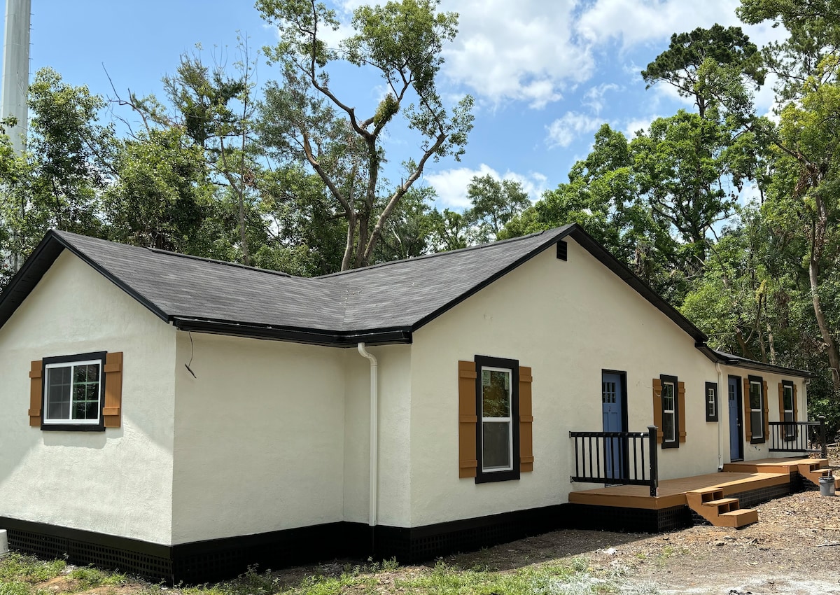 A single-story building showcases a light-colored exterior with black trim and brown shutters on the windows. A blue door leads to the entrance, while a wooden deck extends along the front, surrounded by lush greenery and tree cover.