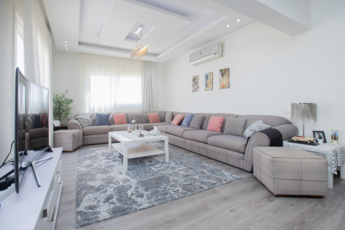 A spacious living area features a large, gray sectional sofa adorned with colorful cushions. A simple coffee table is centered on a patterned area rug, with natural light filtering through sheer curtains. Warm ambiance is enhanced by decorative wall art and a flat-screen television.
