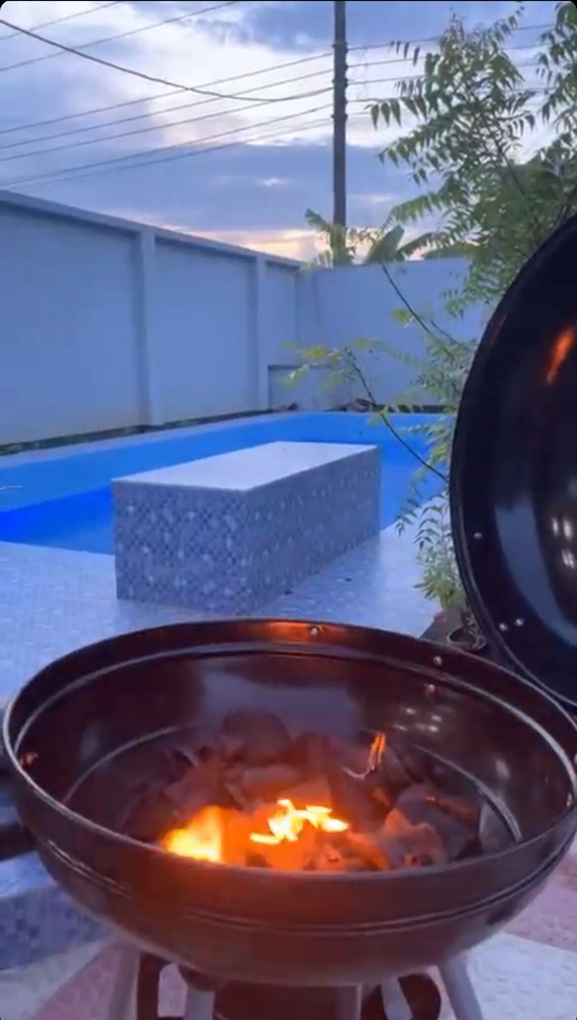 A barbecue grill is positioned prominently in the foreground, with glowing charcoal ignited inside. In the background, a lighted swimming pool is visible, framed by a gray wall and greenery under a twilight sky.