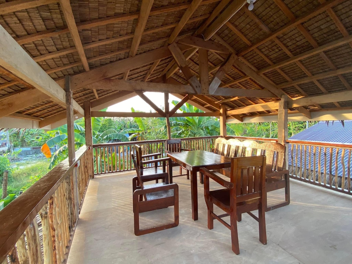 An open-air terrace features wooden furniture, including a large dining table surrounded by several chairs. The ceiling boasts a natural thatched design, and lush greenery is visible in the background, creating a peaceful outdoor setting.