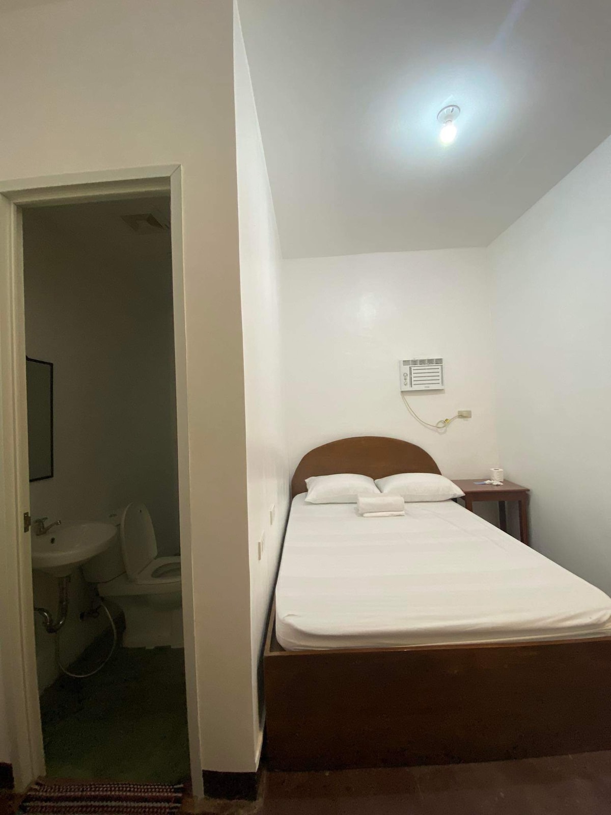 A simple room is depicted, featuring a single spacious bed with white linens and folded towels. A small bedside table is positioned beside the bed. A bathroom door is visible, indicating access to a private restroom. The walls are painted white, contributing to a clean and bright appearance.