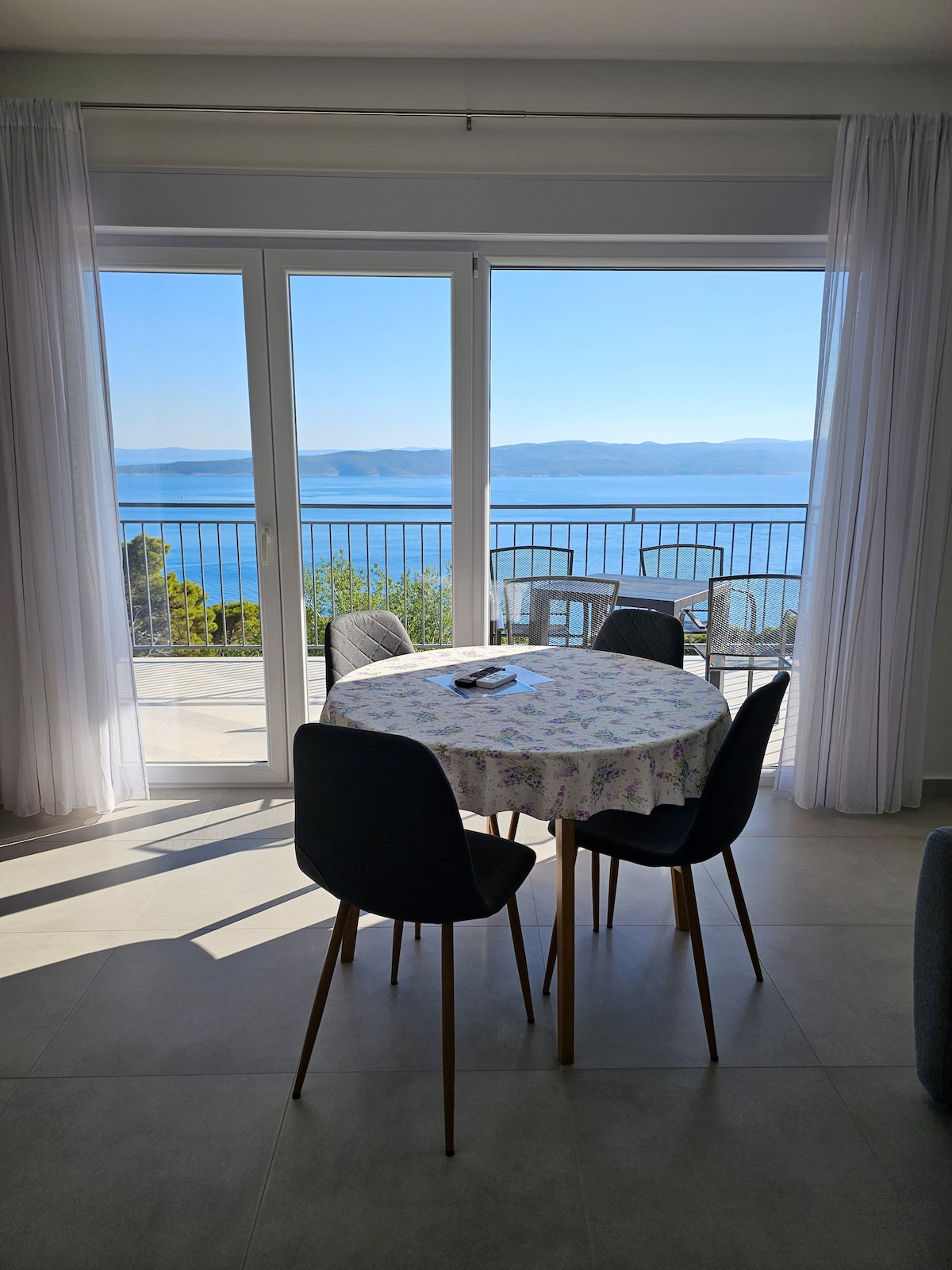 A round dining table with a floral tablecloth is positioned near expansive glass doors, offering a clear view of the sea and islands beyond. Tall chairs surround the table, and light drapes flow gently beside the doors, enhancing the bright and open ambiance.