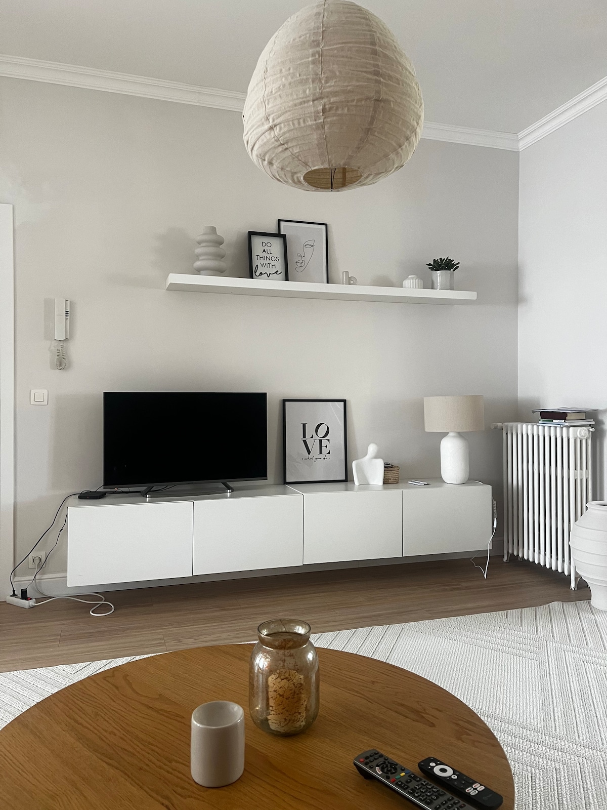 The living area features a light-toned wall and a wooden coffee table at the forefront. A white media console holds a television and decorative items. Above, a shelf displays framed art and a small plant. A pendant light hangs from the ceiling, providing illumination.