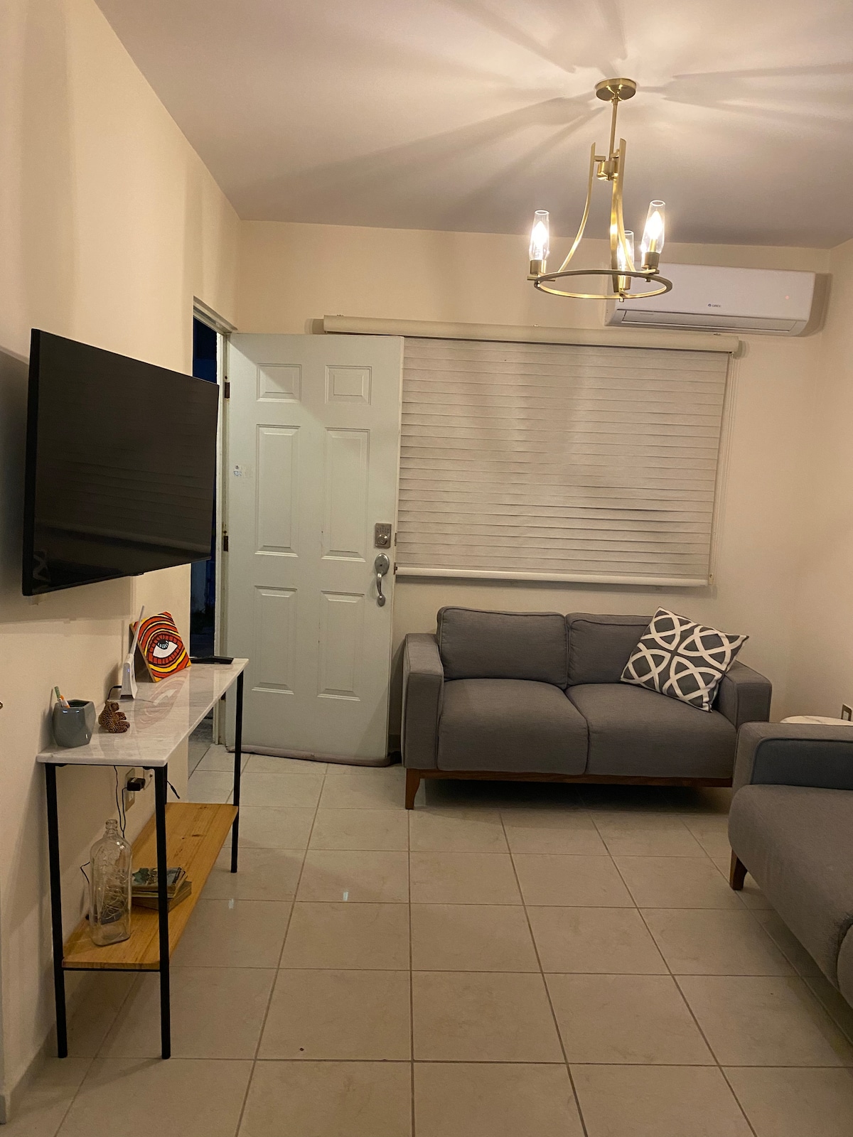 A well-lit living area features a gray sofa adorned with decorative cushions. A wall-mounted television is positioned above a small console table, complemented by a light fixture hanging from the ceiling. The entrance door is visible, along with a window covered by a light blind.