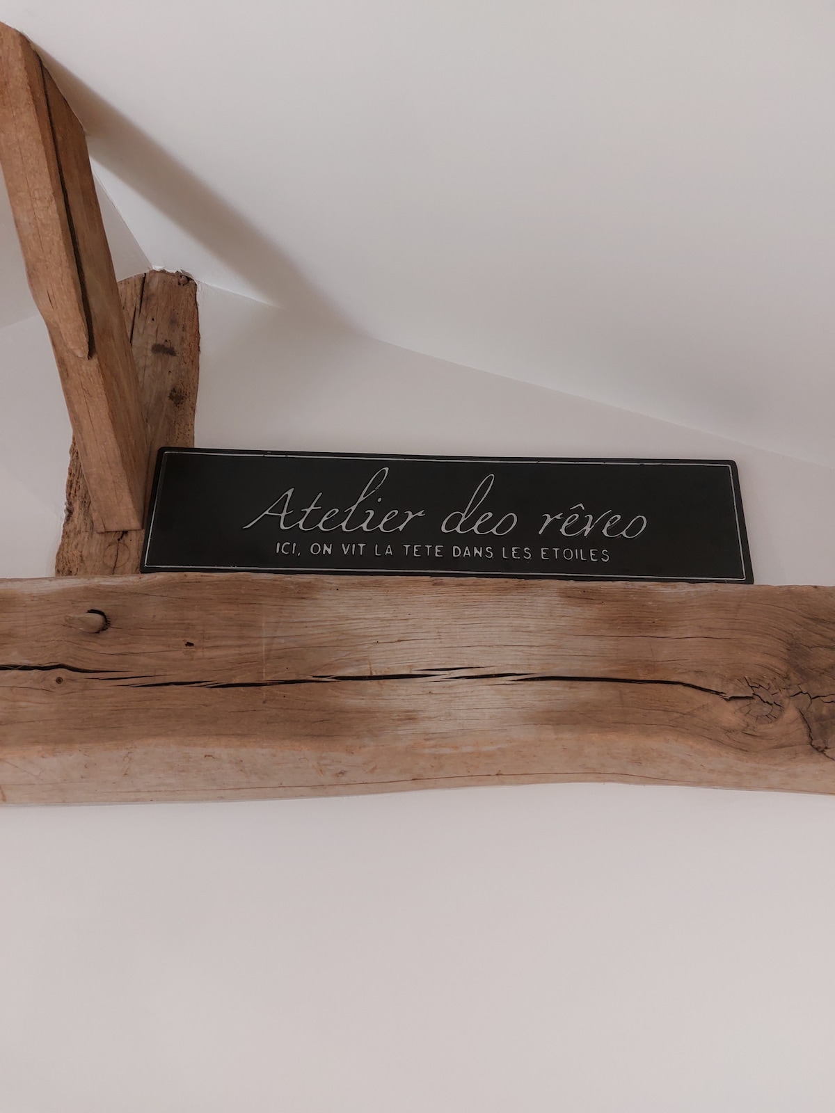 A rustic wooden beam features a black sign with white lettering that reads 'Atelier deo rêveo' and a phrase below. The soft textures of the wood are contrasted by the smooth surface of the sign, contributing to a charming, artistic atmosphere.