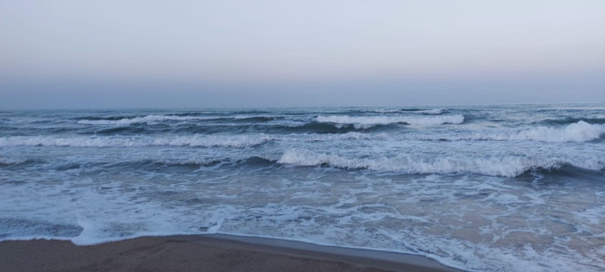 The image captures a serene beach scene at dawn, showcasing gentle waves lapping against the shore under a soft sky gradient. The horizon meets the ocean, creating a tranquil atmosphere with subtle variations in color from light blue to soft gray.
