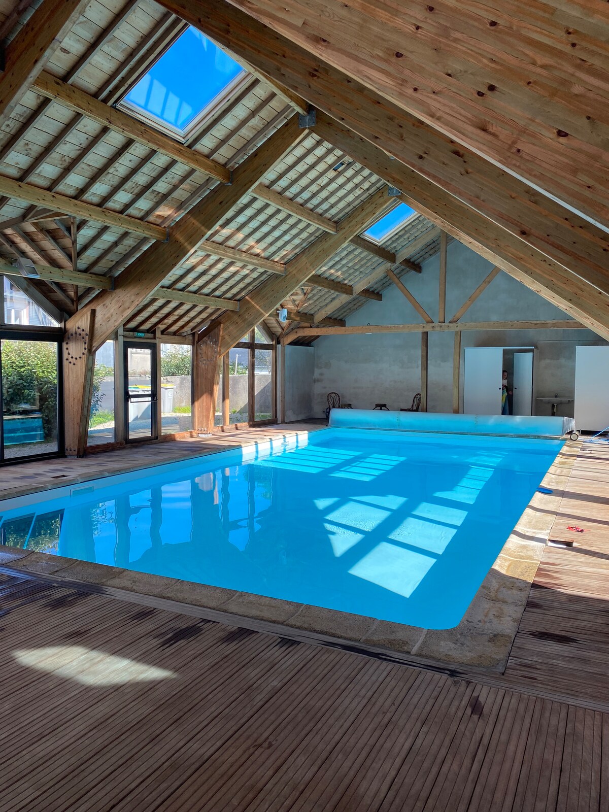 An indoor heated pool is featured within a spacious wooden structure, showcasing high ceilings with exposed beams. Large skylights allow natural light to illuminate the clear blue water. Floor-to-ceiling windows provide views of the outdoor area, enhancing the atmosphere.